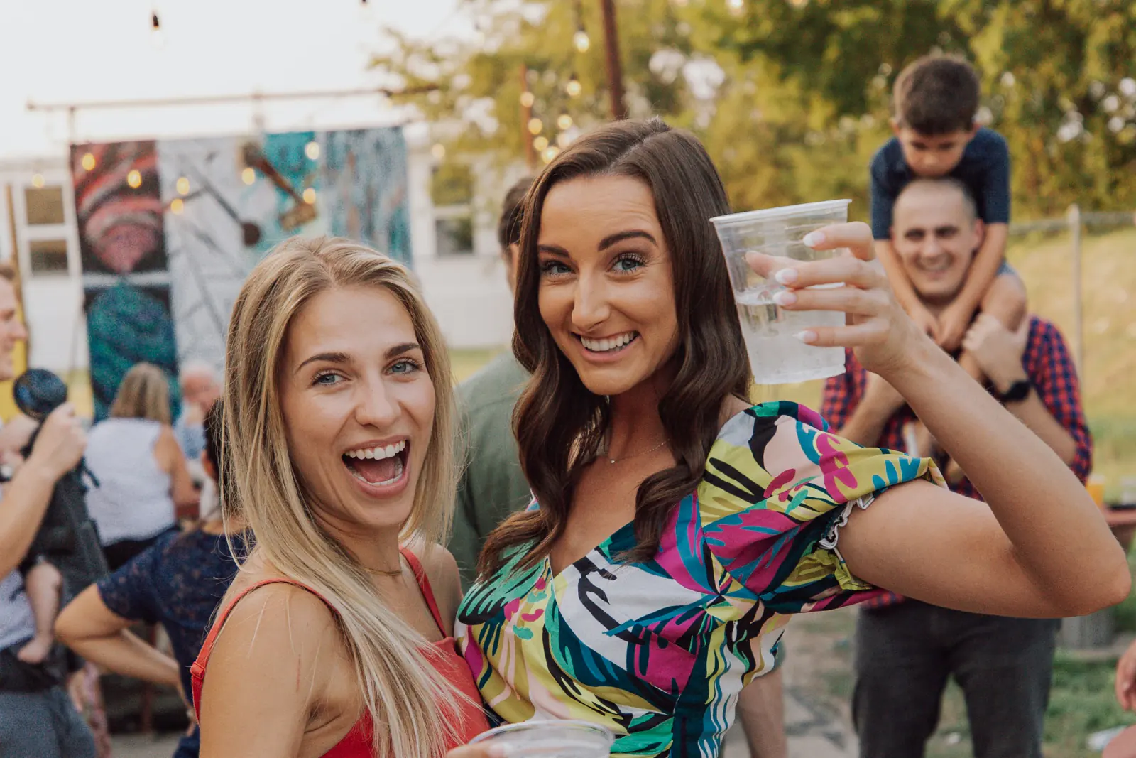 Two friends laughing and raising their cups at an outdoor party under string lights